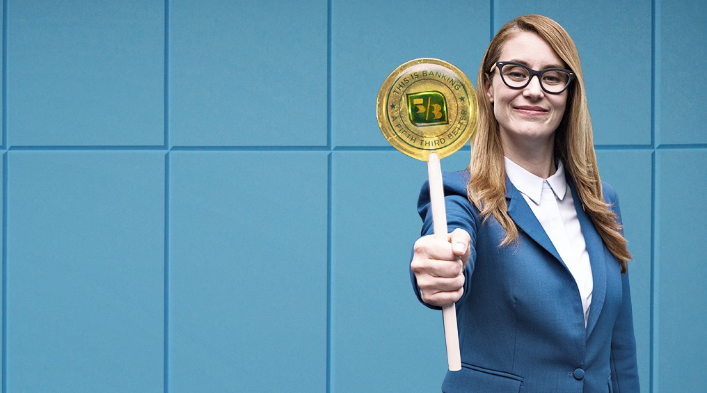 Woman in front of blue wall holding big yellow lollipop with Fifth Third logo on it.