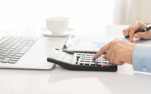 Man using a calculator next to a laptop and a cup with a saucer.