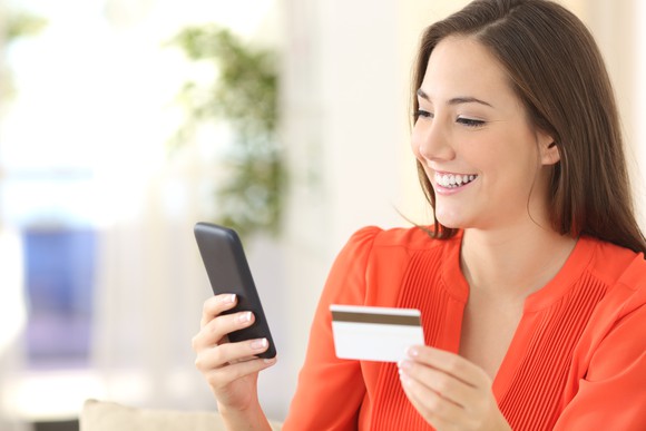 Woman in a red blouse smiles at her phone while holding a credit card in her other hand.