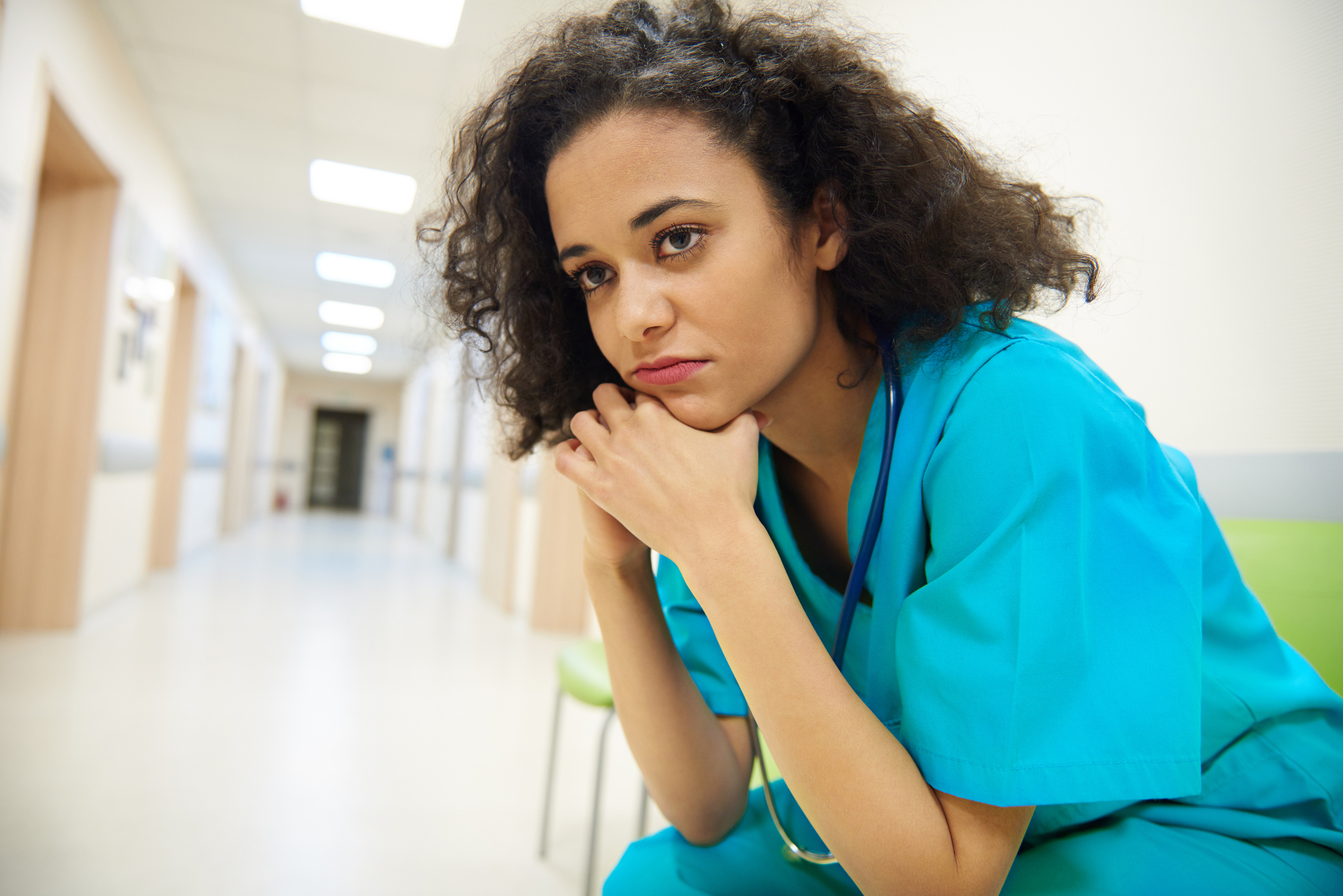 A worried nurse sitting in a hallway with her head resting on her hands.