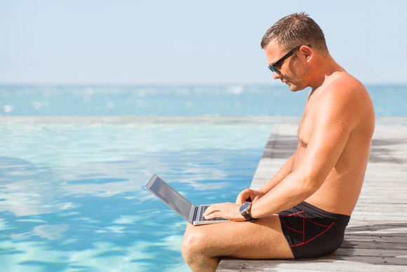 Man in swim trunks using a laptop by a pool