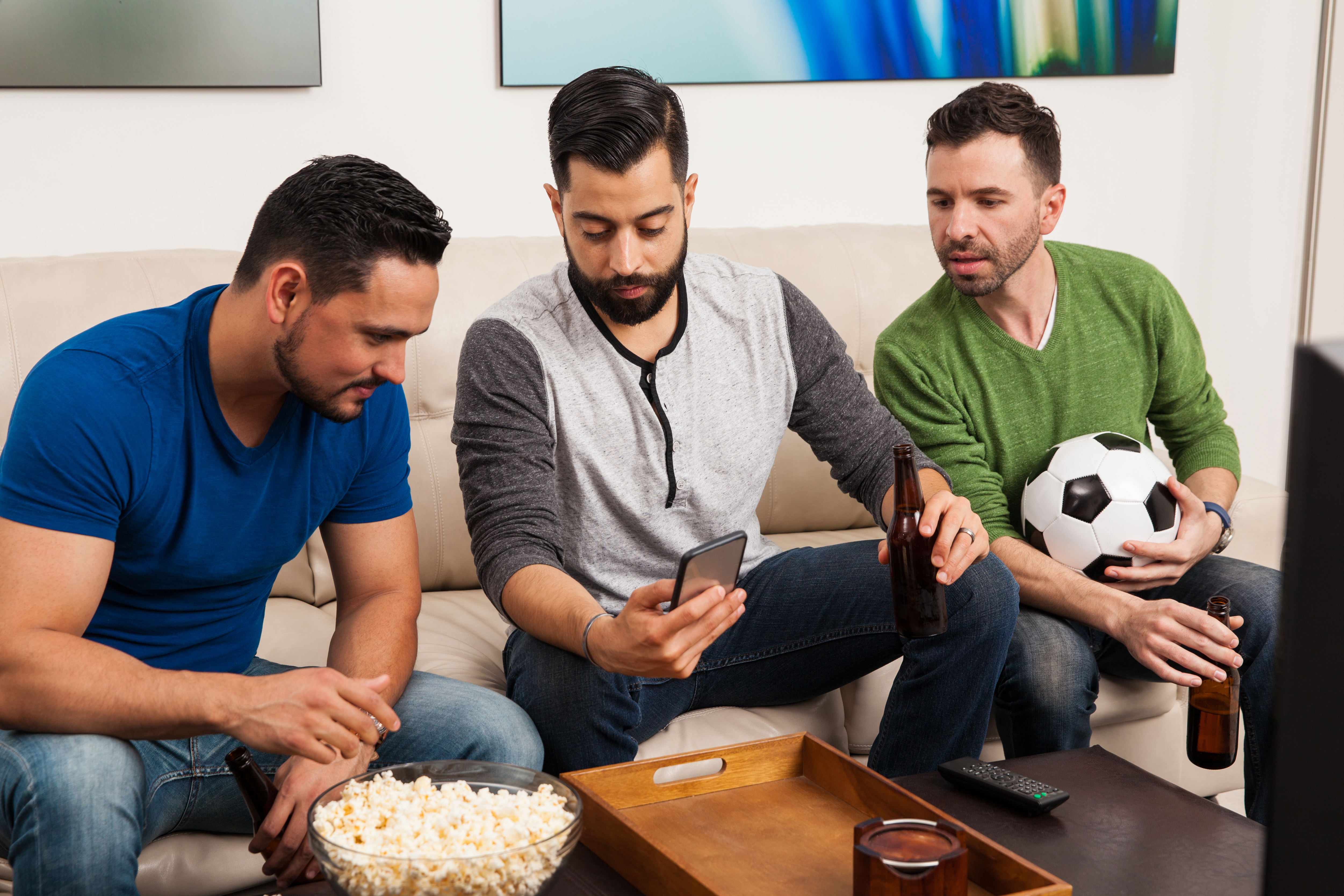 Three people sitting on a couch looking at a smartphone with one person holding a soccer ball and a bowl of popcorn on the table in front of them.