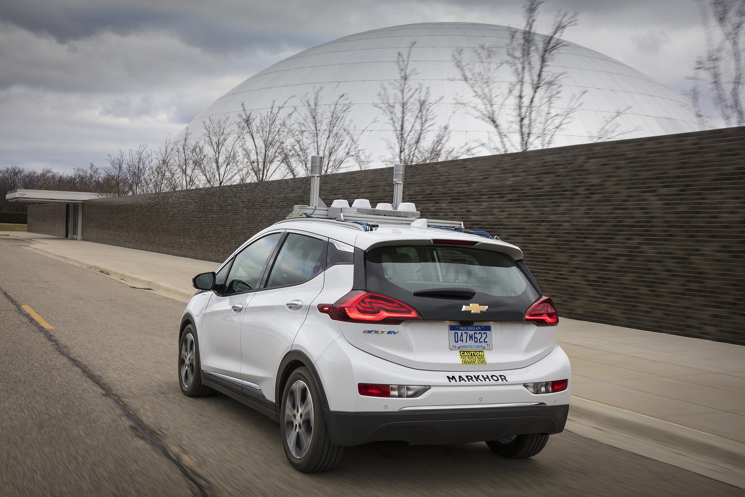 White car on a road with autonomous driving sensors on top of it, in front of a large dome.