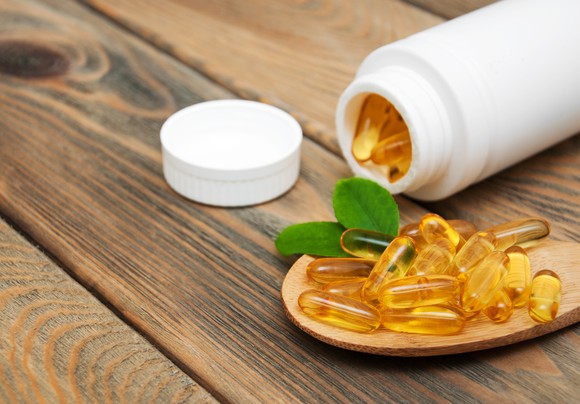 A white bottle containing softgel capsules tipped over, with softgels lying on a spoon adjacent to the bottle.