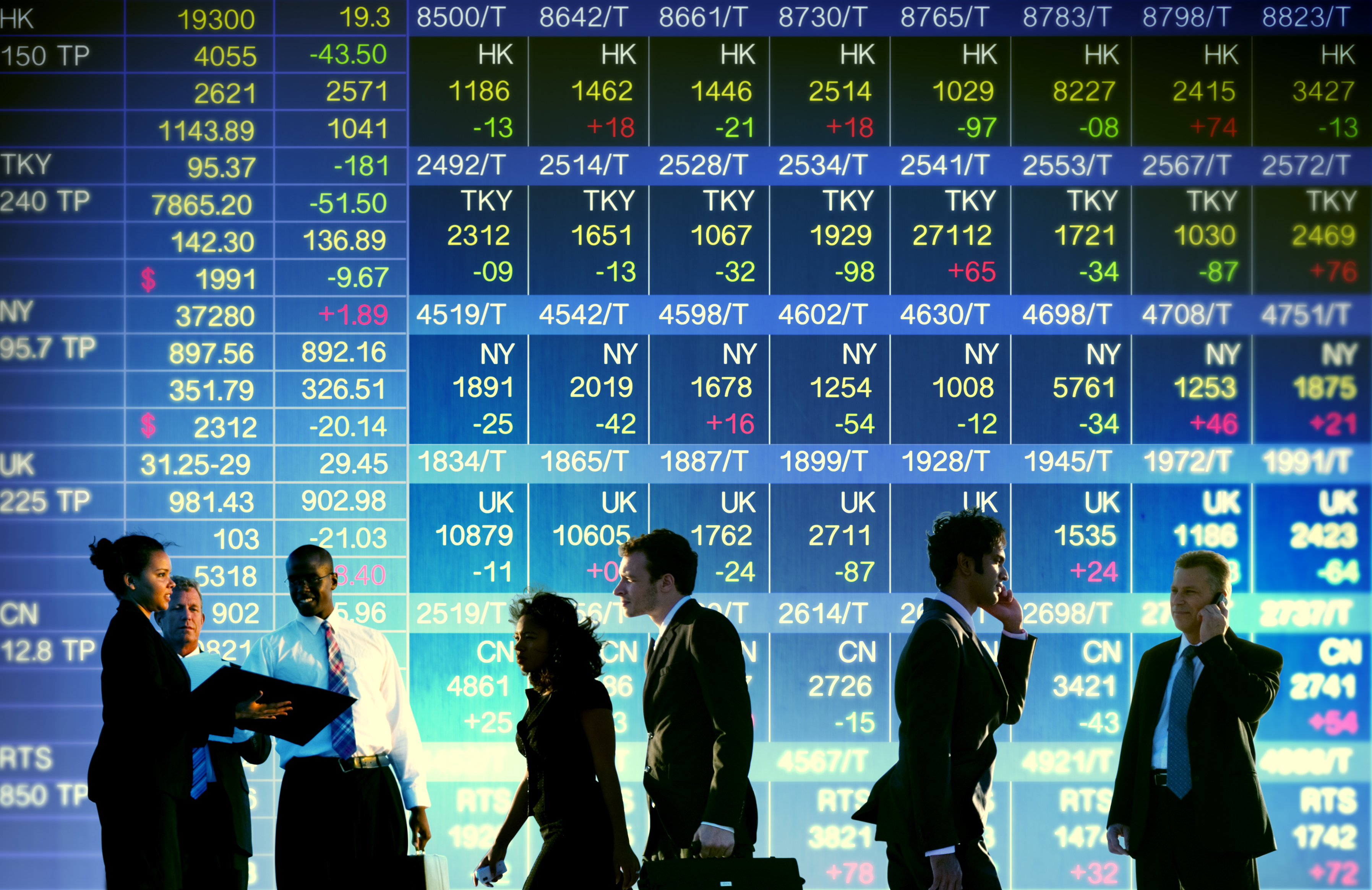 Men and women in suits, standing and walking in front of a display of stock prices.