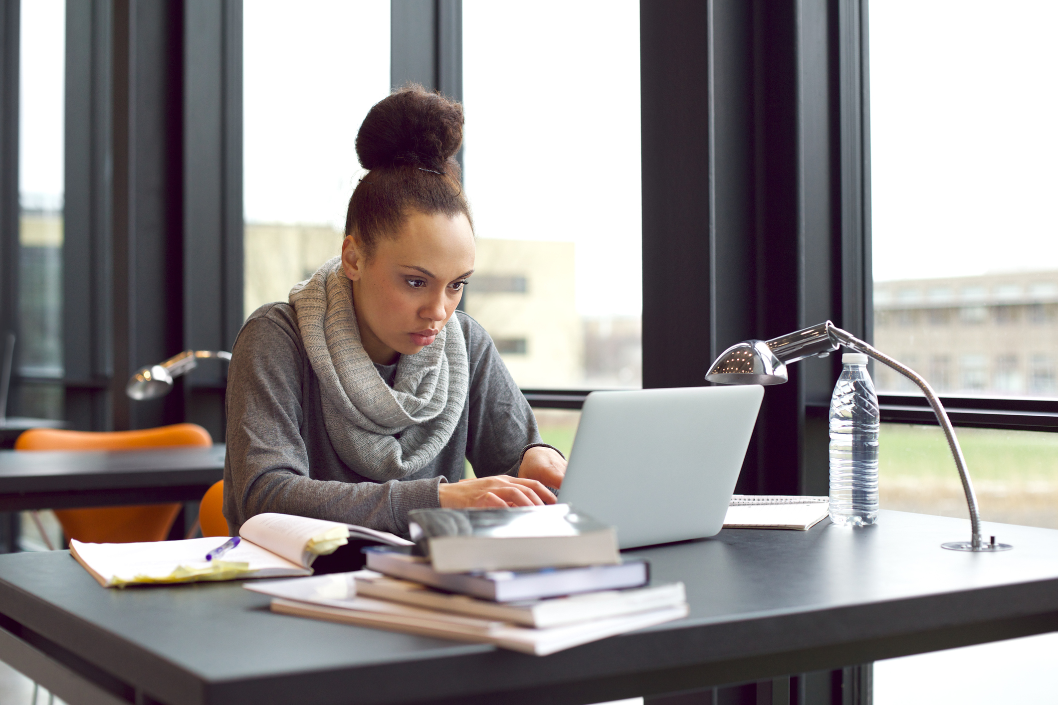 Young female at a laptop next to a pile of books and open notebook