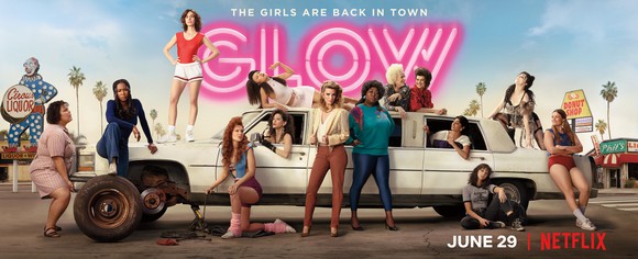 Cast members from season two of the Netflix original series GLOW (Glorious Ladies of Wrestling) posing around a car with the front tire off.