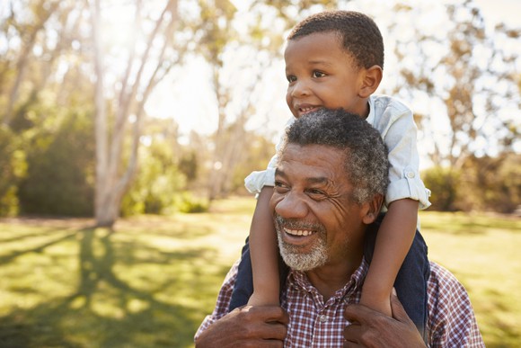 A grandparent carrying his grandson on his shoulders through a park.