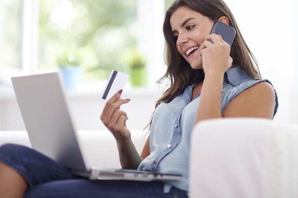 A woman talking on her smartphone with her laptop on her lap and a credit card in her right hand.