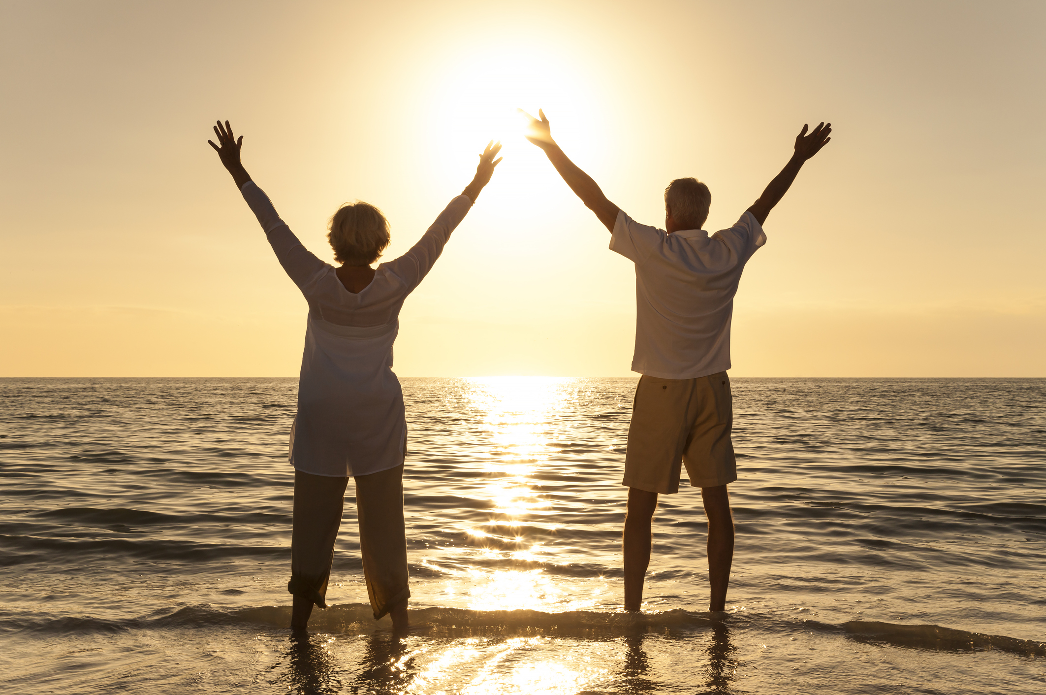Senior woman and man on a beach at sunset with their arms raised high