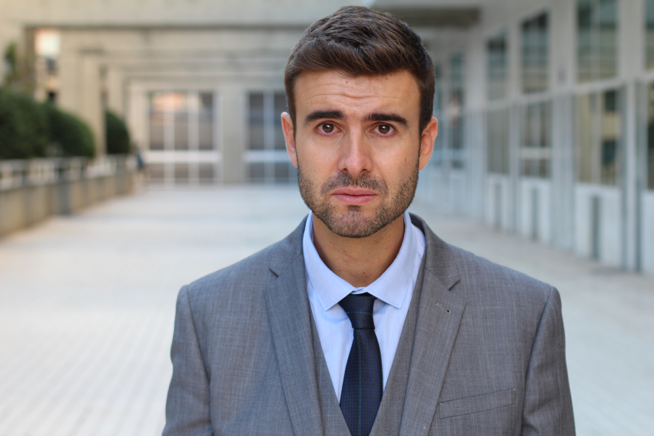 Man in suit with sad expression standing in a long, empty hallway.