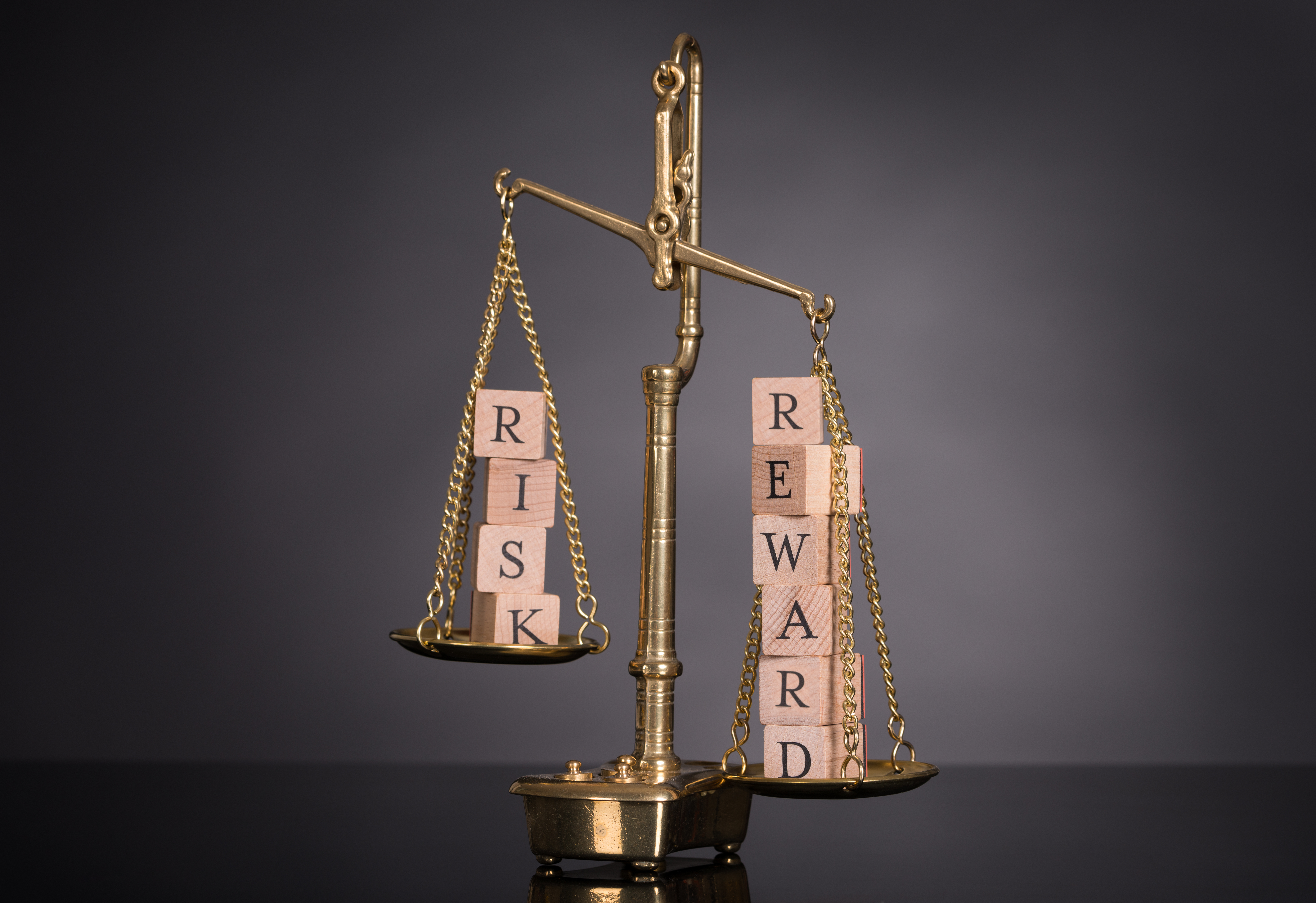 Balance scales with blocks spelling Risk on one side and Reward on the other.