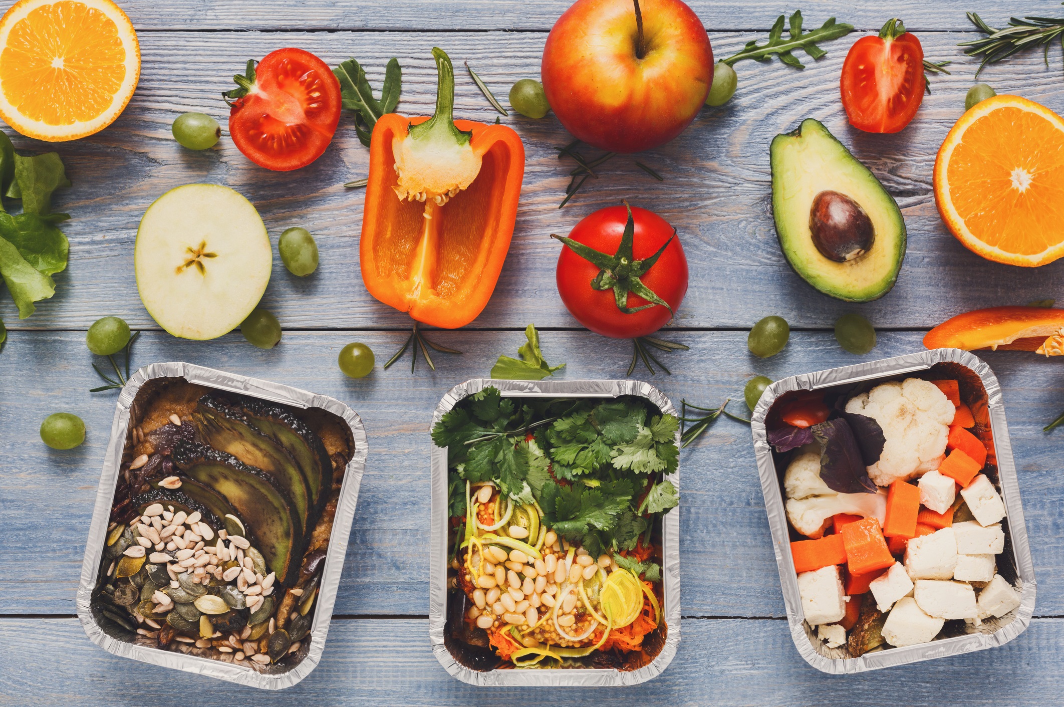 Wooden table covered with fresh fruits, vegetables and spices. Three assembled meals in aluminum trays show combined fresh ingredients.