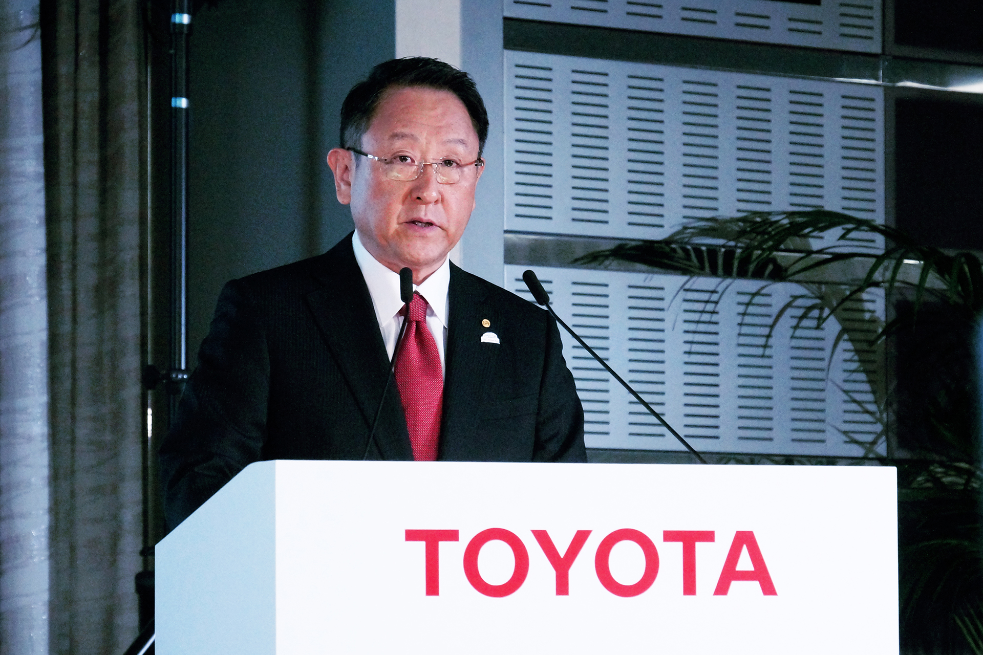 Toyoda speaking at a white podium with "TOYOTA" in red on the front.