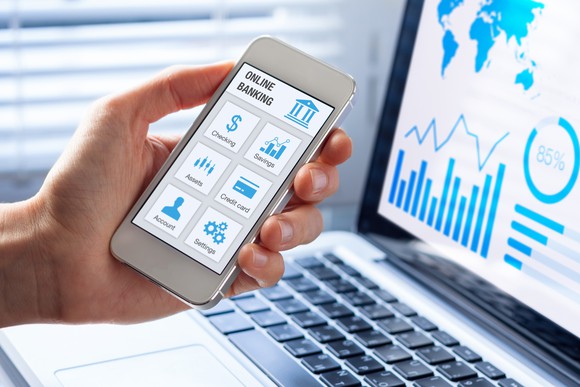 Hand holding a smartphone displaying an online banking app, computer with banking data in the background