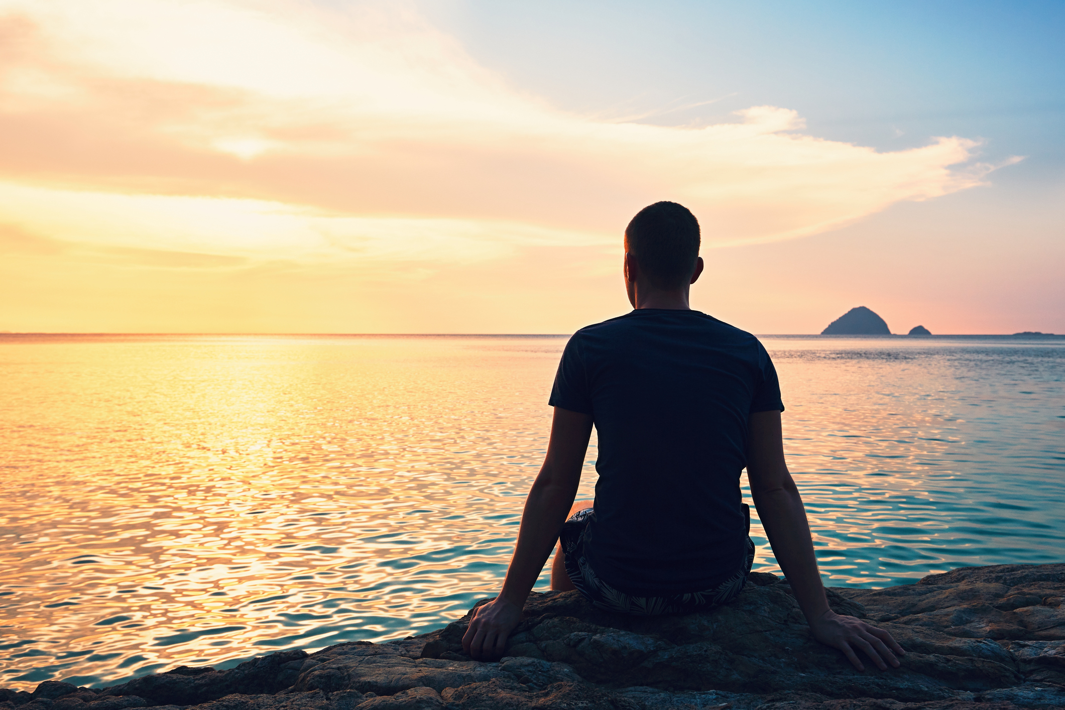 Man sitting facing the water at sunset