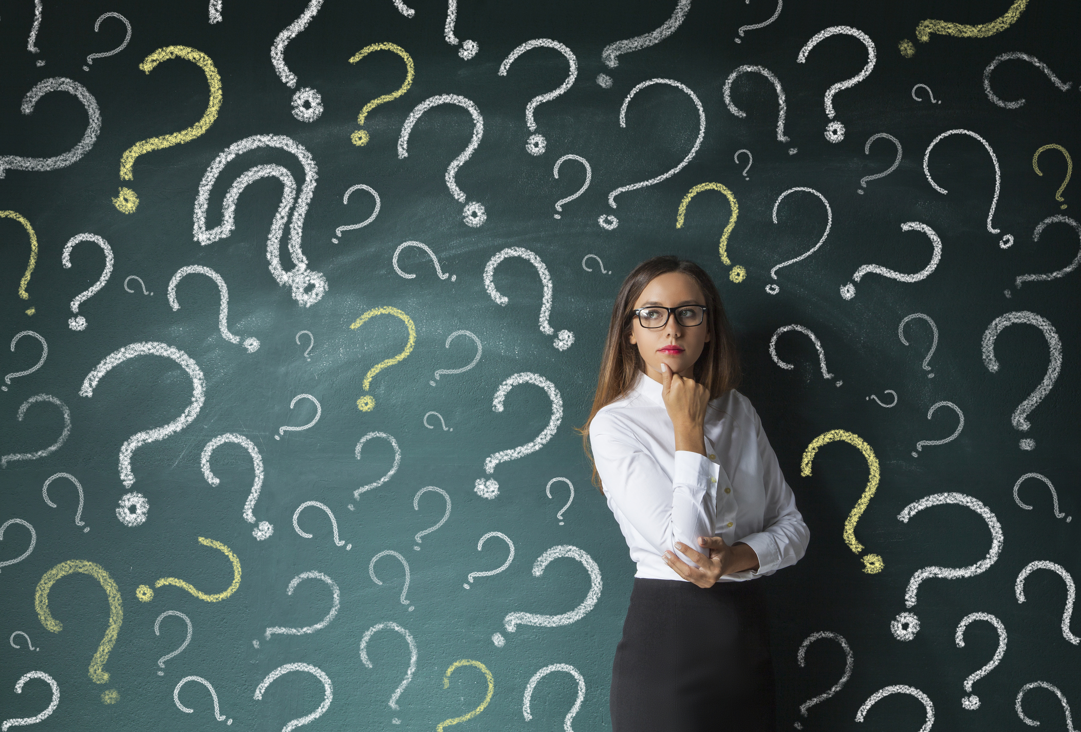 A businesswoman standing in front of a chalkboard with many question marks drawn on it.