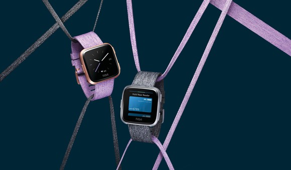 Two versions of the Fitbit Versa hanging from strings.