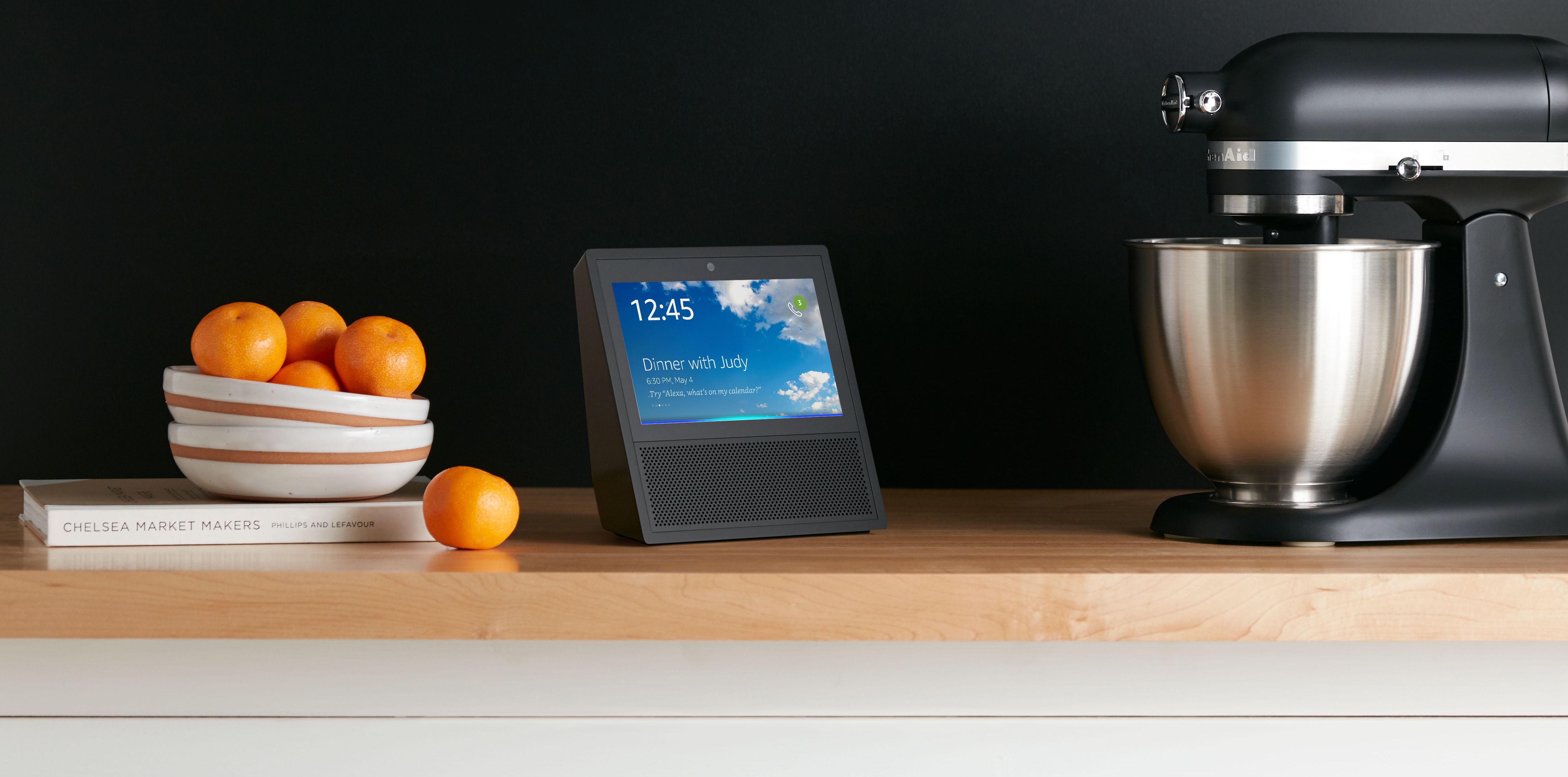 Echo Show on a kitchen counter next to a stand mixer and a bowl of oranges