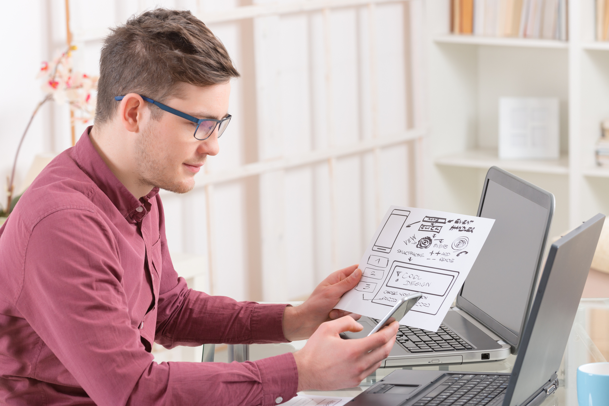 A man sits at a laptop holding a piece of paper detailing plans.