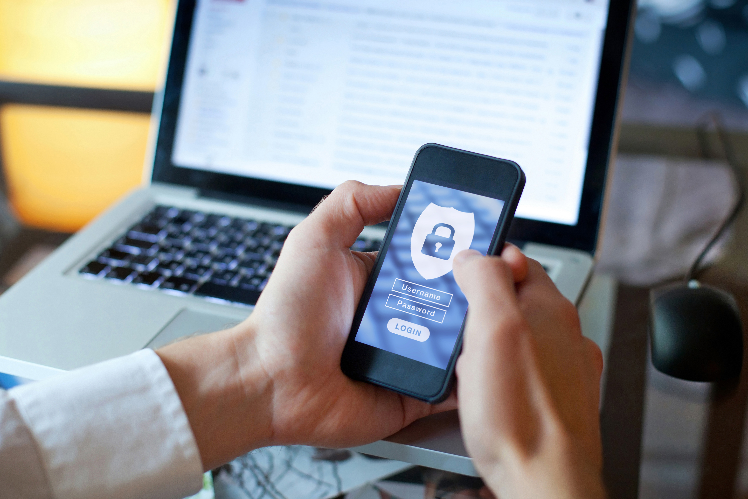 Person with open laptop holding a smartphone with a security icon and login screen