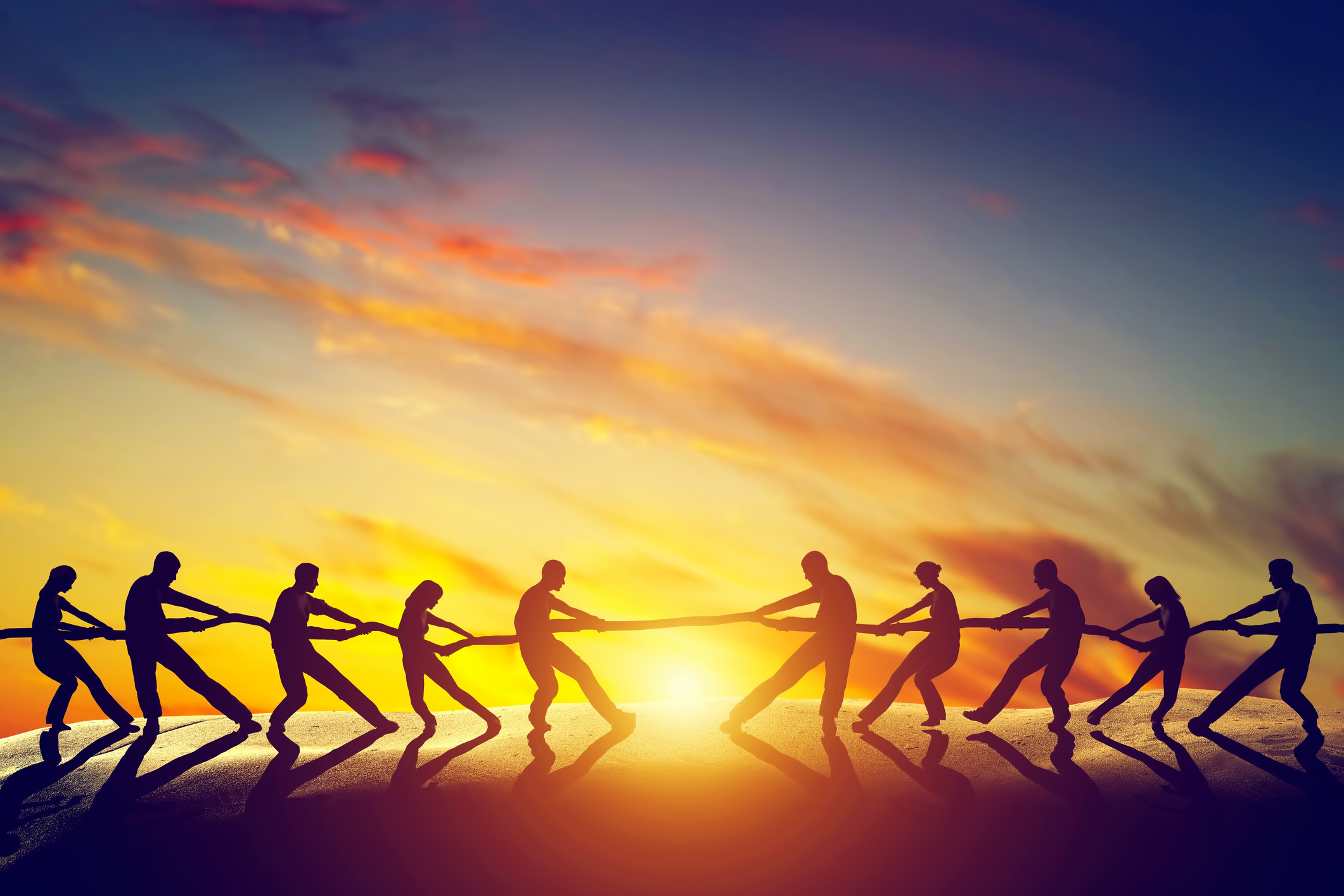 A group of people in silhouette playing tug of war during sunset.