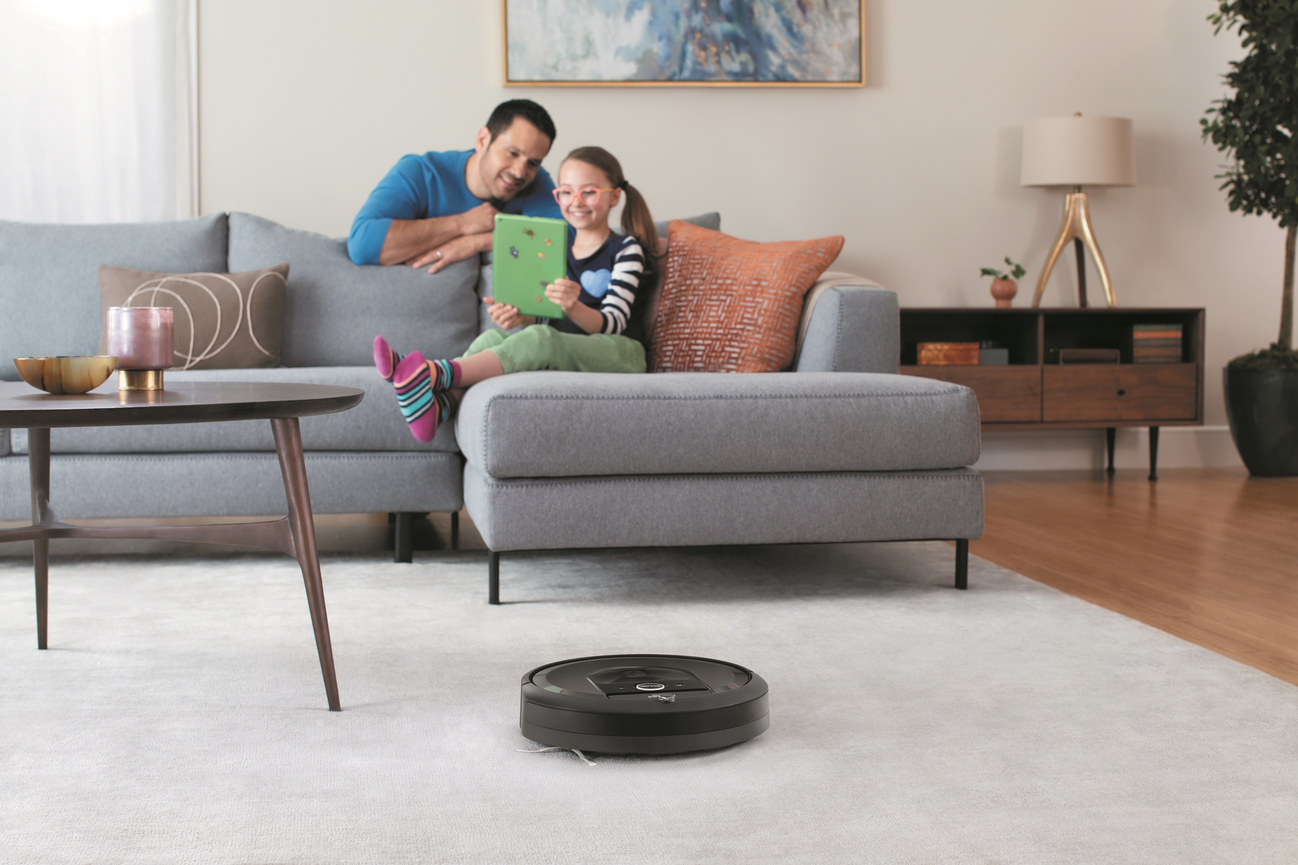 A young girl sitting on a couch looking at a tablet with her father looking over her shoulder, as a robotic vacuum cleans a carpet in the foreground.