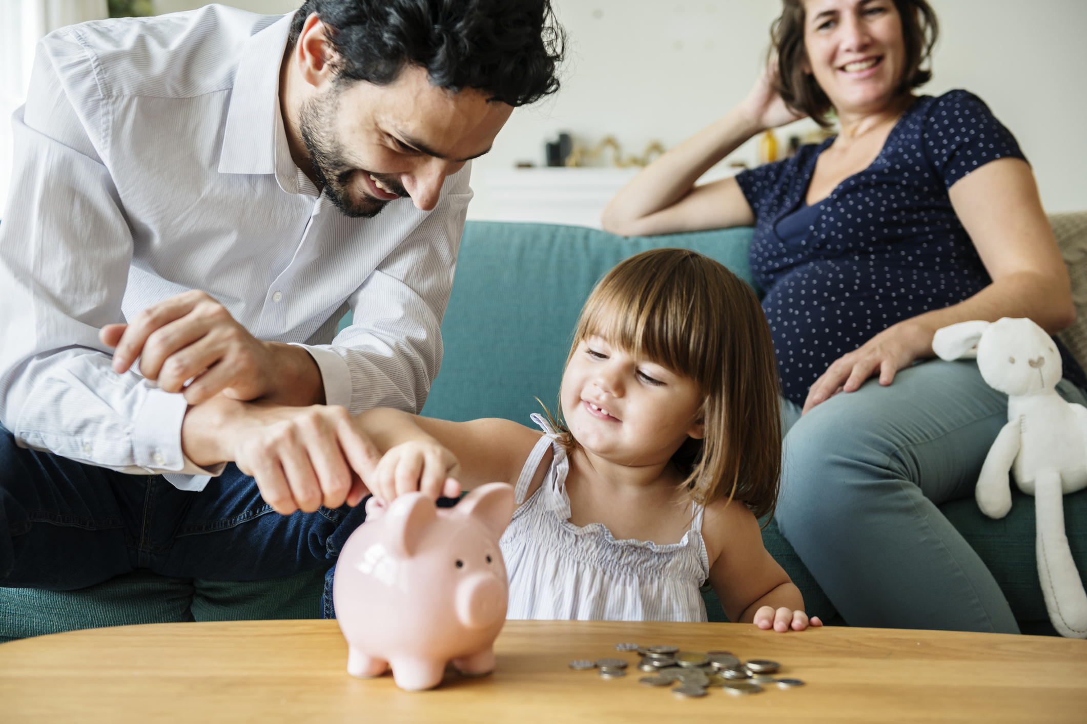 Young girl inserts coin into piggy bank while her father and pregnant mother look on.