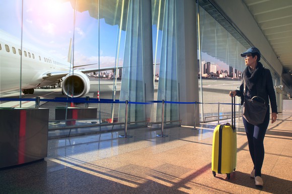 A female traveler in an airport terminal with luggage stares at a plane through large glass window.
