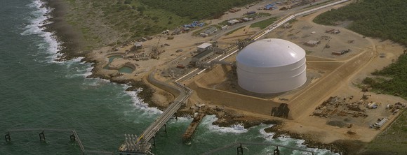 Beach-side facility including a large white storage tank and various lines heading out a pier toward a dock.