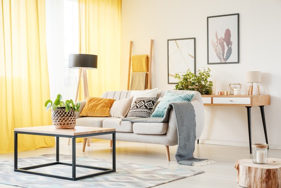 A living room that includes a plant on a table, a lamp, yellow curtains, framed art on the wall, and pillows on a sofa