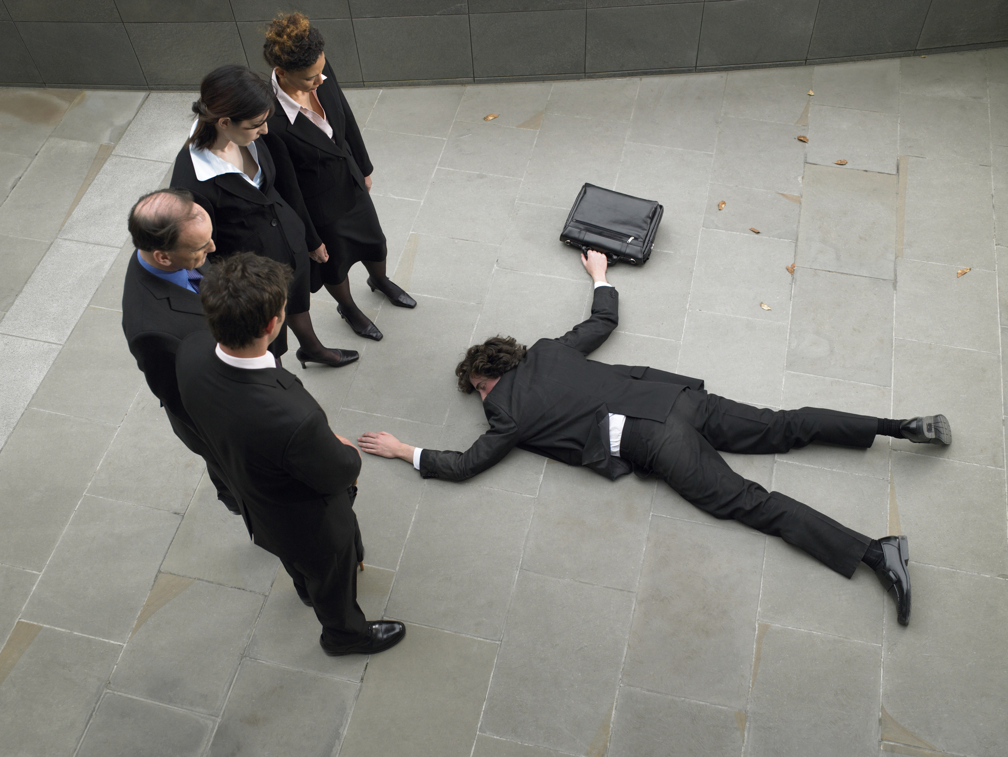 A man wearing a suit and holding a briefcase lies face down on a concrete surface, as if knocked down, while four other people in suits look down at him. 