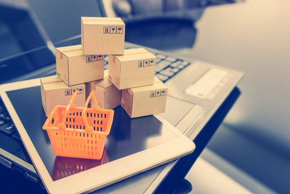 A pile of miniature boxes and a miniature shopping basket on top of a tablet and laptop.