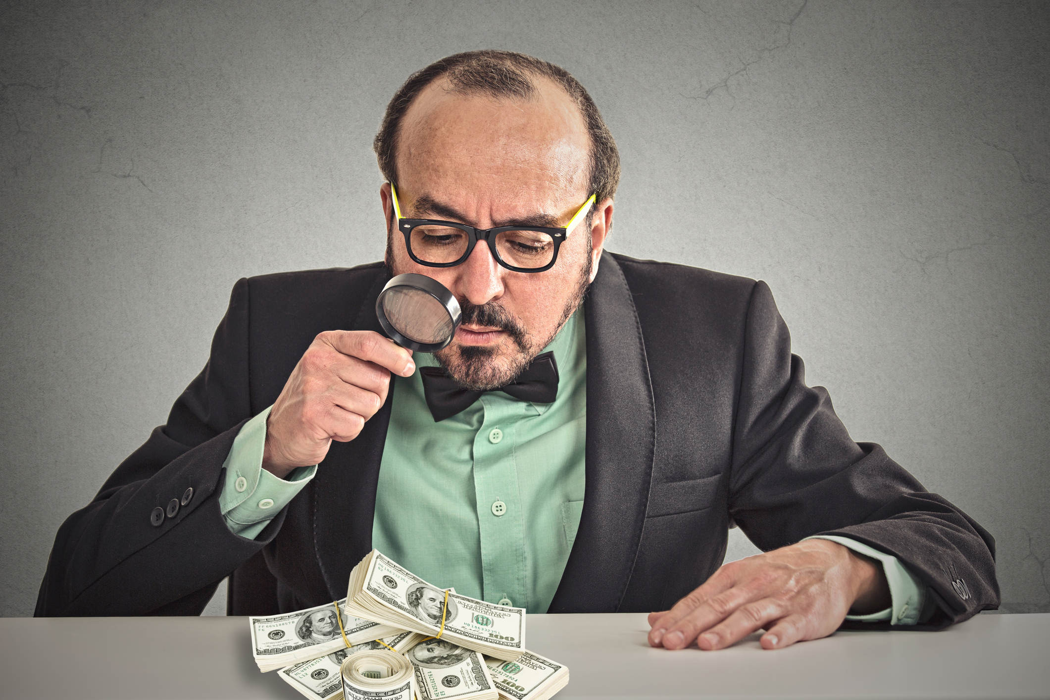 A businessman in a suit looking at a pile of money with a magnifying glass