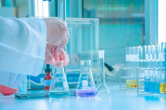 A lab worker's hands holding a beaker with some liquid in it that's sitting on a table along with other beakers and test tubes