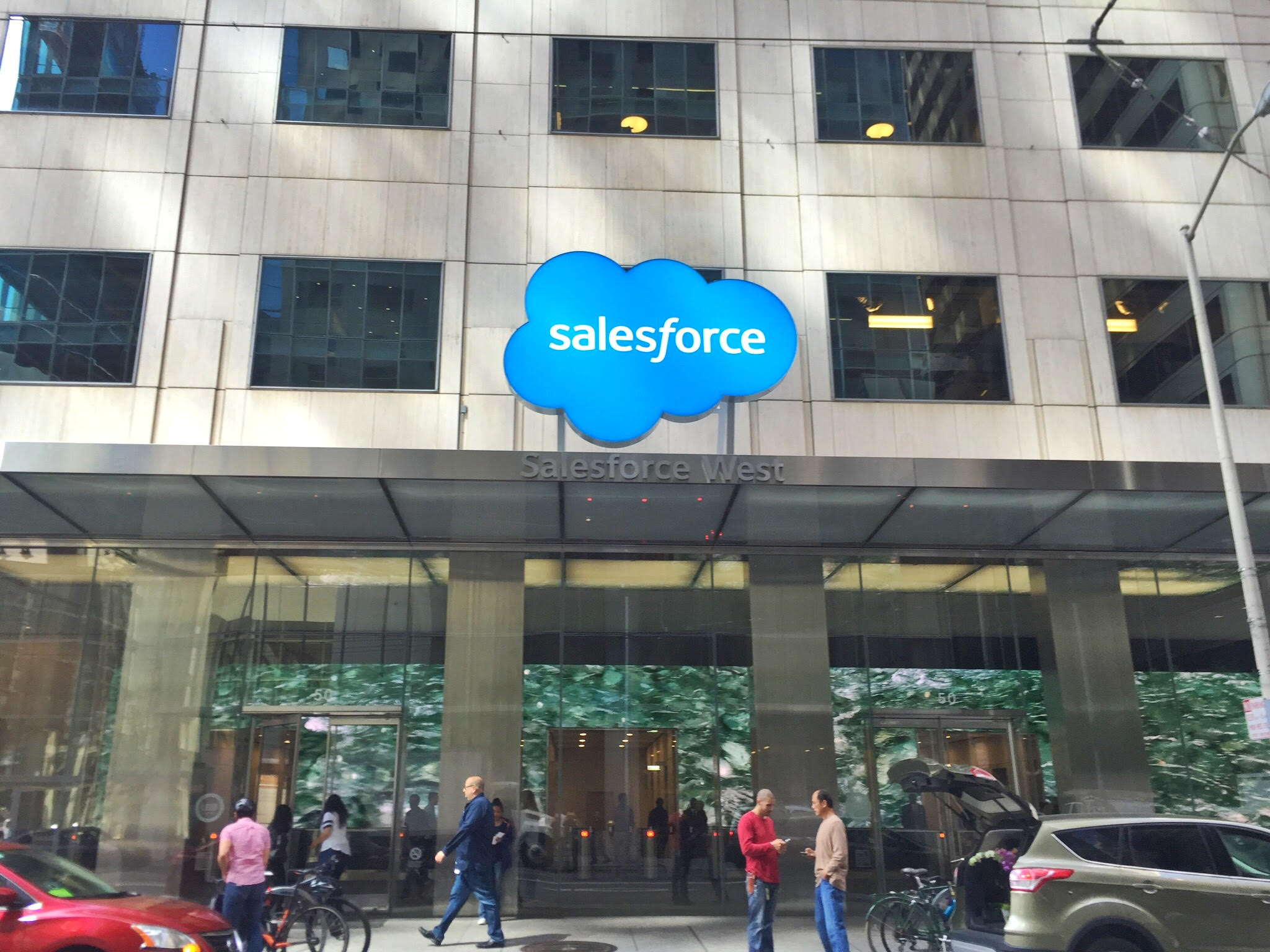 People walking about and standing in front of a large building with the Salesforce logo over the entrance.