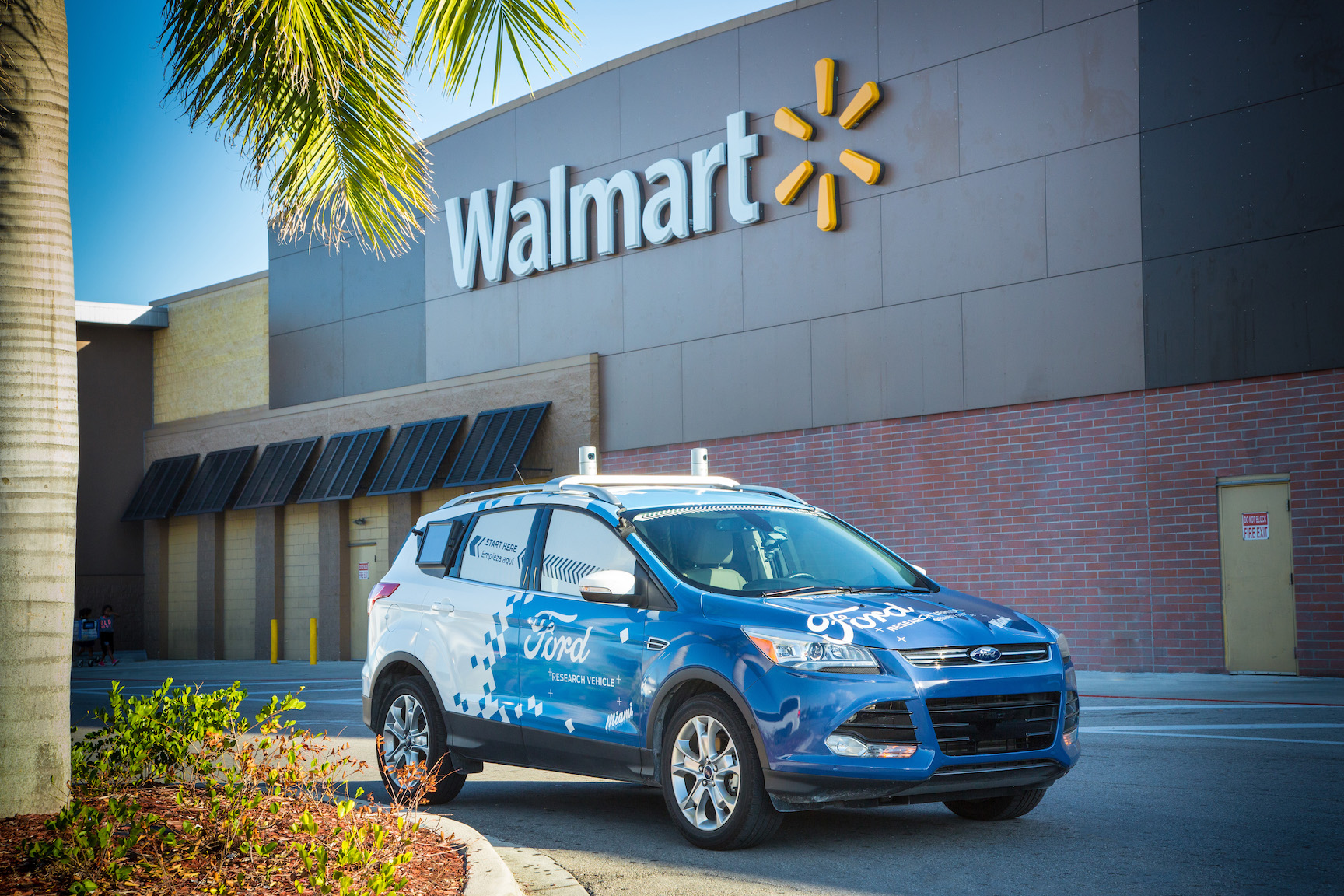 A Ford Escape, a compact SUV, with blue and white Ford graphics and visible self-driving sensor hardware, in front of a Walmart store