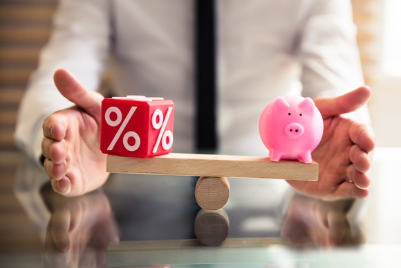 A wooden balance with a die showing a percentage mark on one side and a piggy bank on the other side.