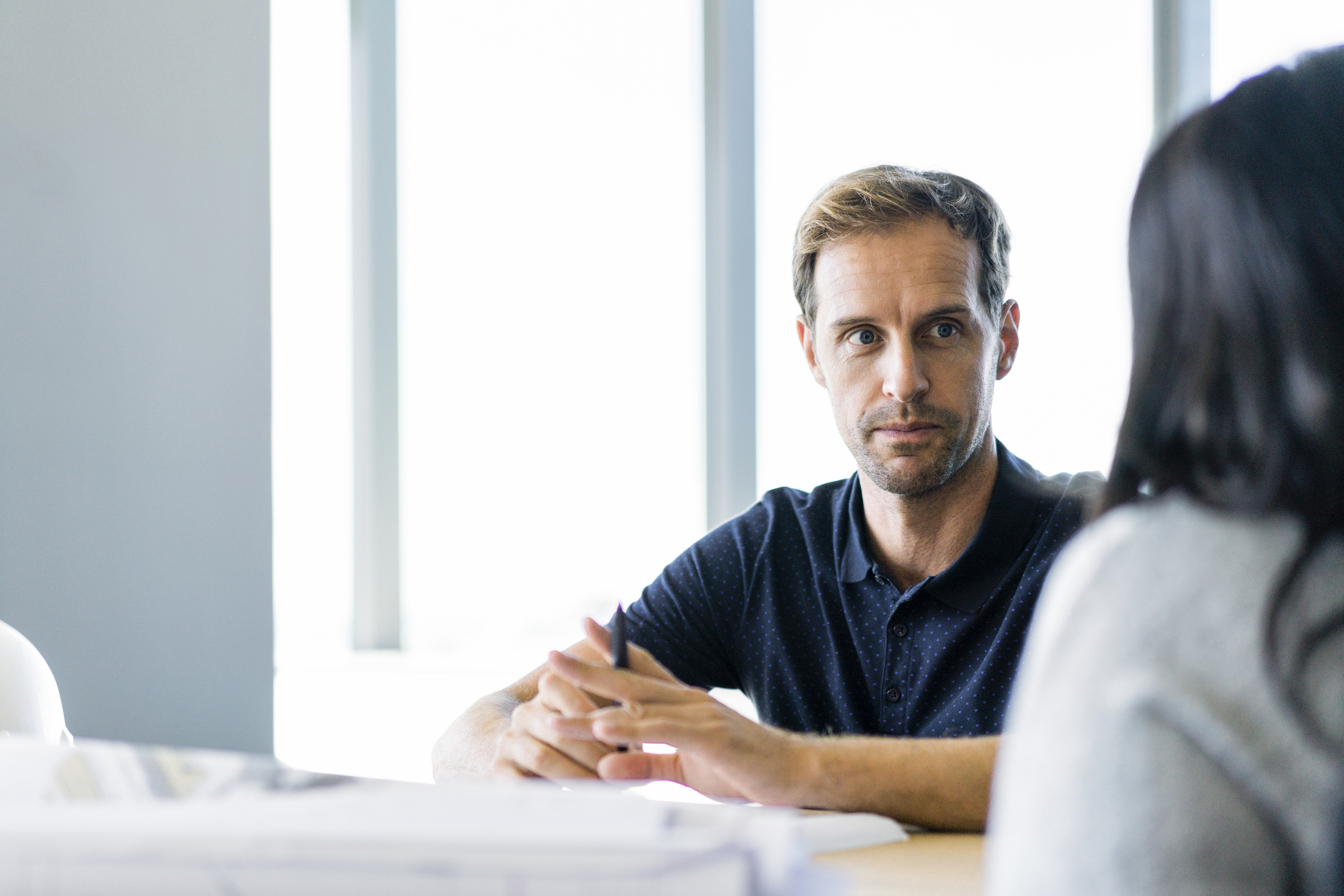 Man with a serious expression and a collared shirt sitting across from a woman in an office