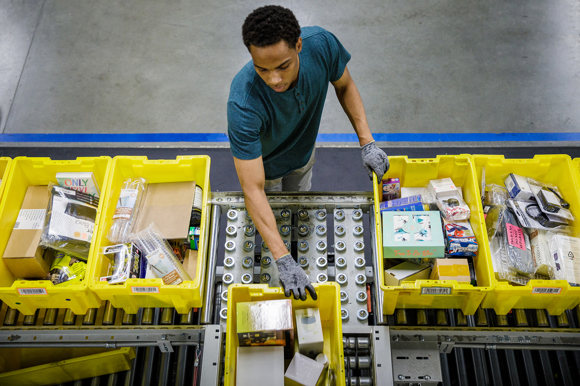 Amazon.com warehouse employee, seen from overhead, sorts yellow bins full of merchandise on a conveyor belt.