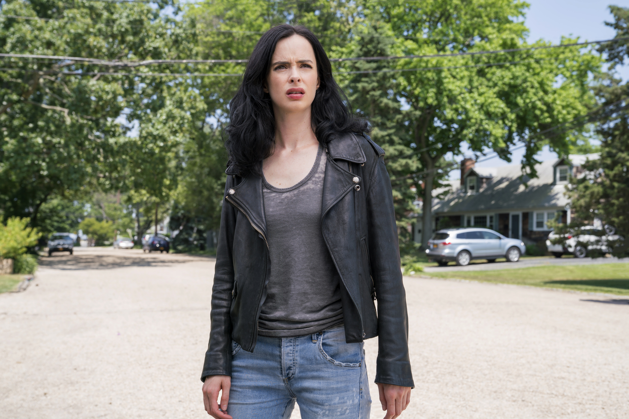 A woman in jeans and a black leather jacket stands in the street looking bewildered.