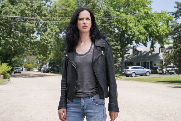 A woman in jeans and a black leather jacket stands in the street looking bewildered.