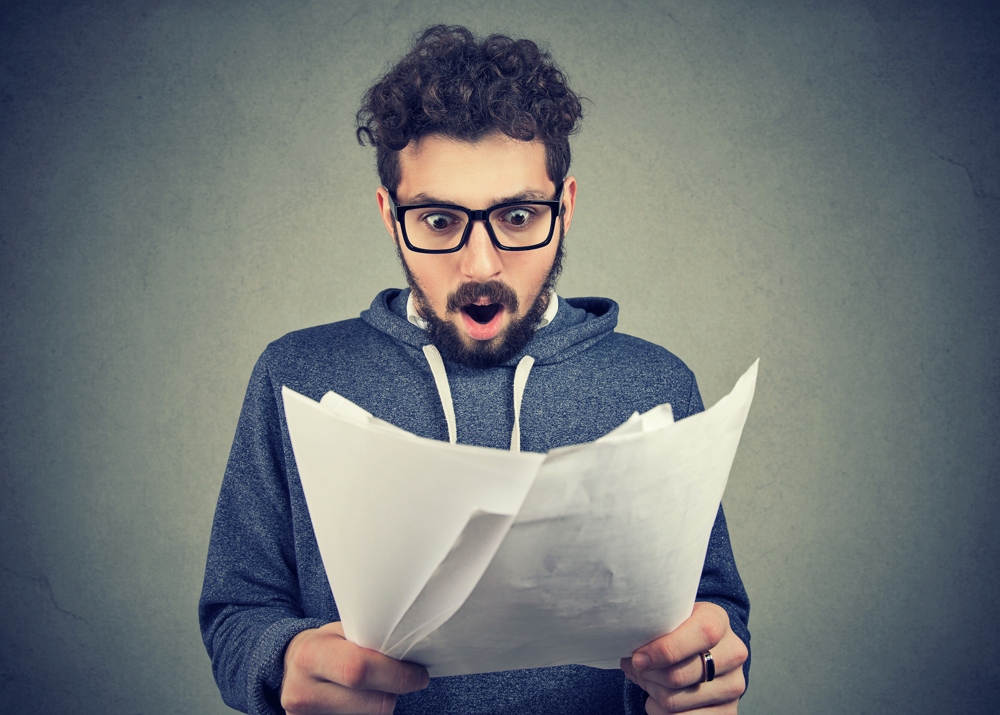 A bearded young man holding and looking at some papers, seems amazed, with his mouth open.