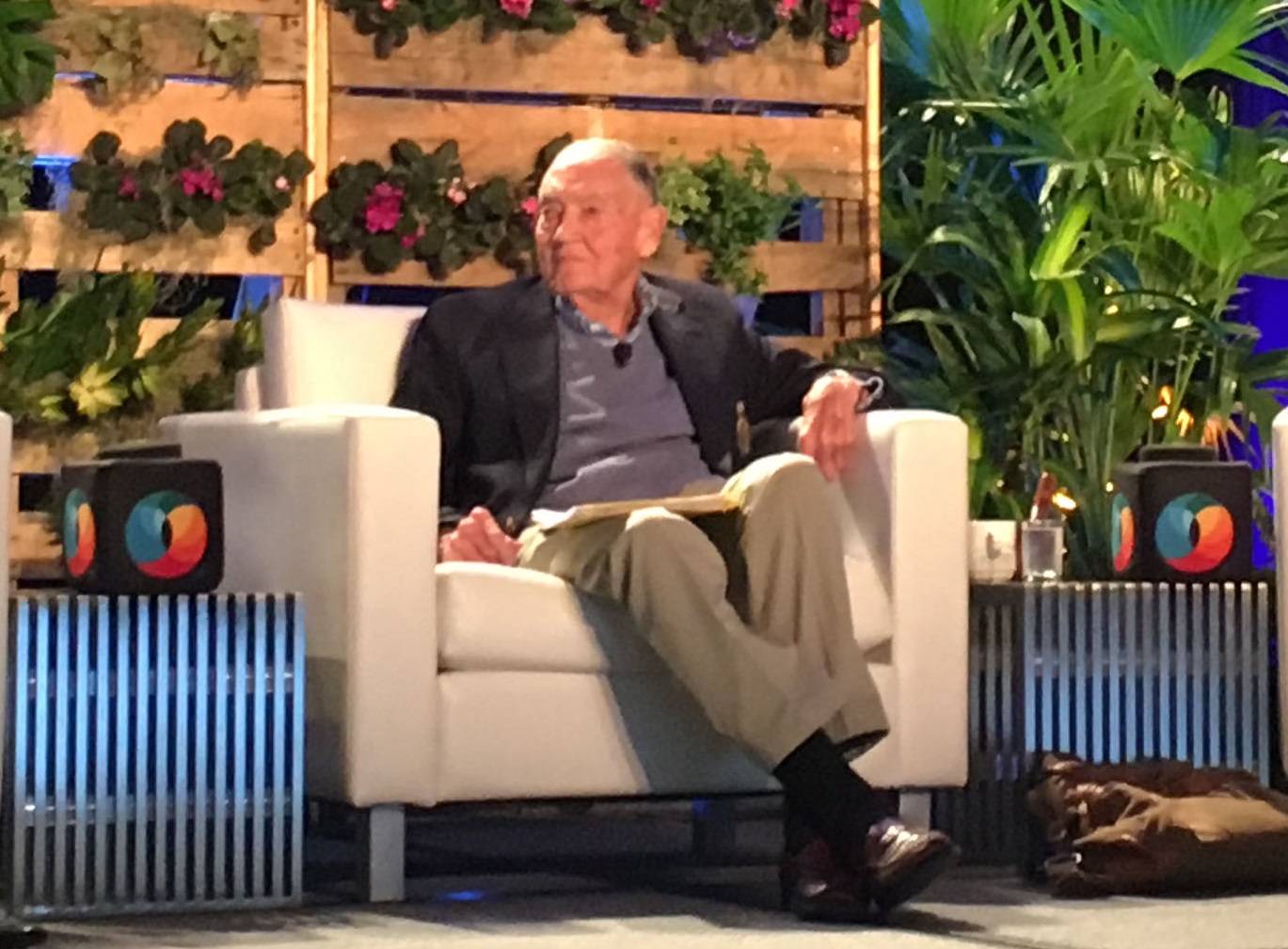 Jack Bogle sitting in a white chair on a stage in front of greenery.