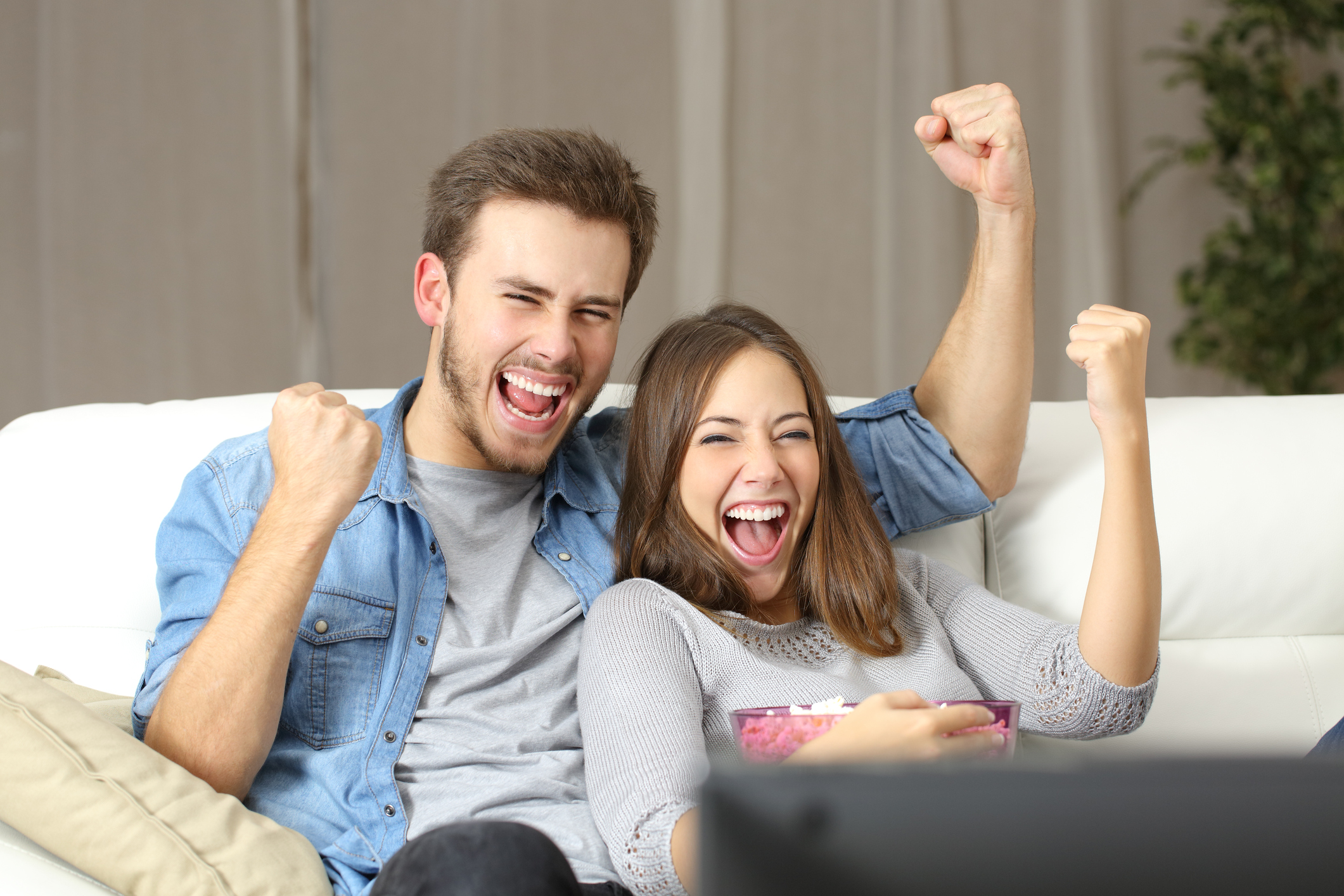 A young couple fist-pumping on the TV couch, cradling a large bowl of popcorn