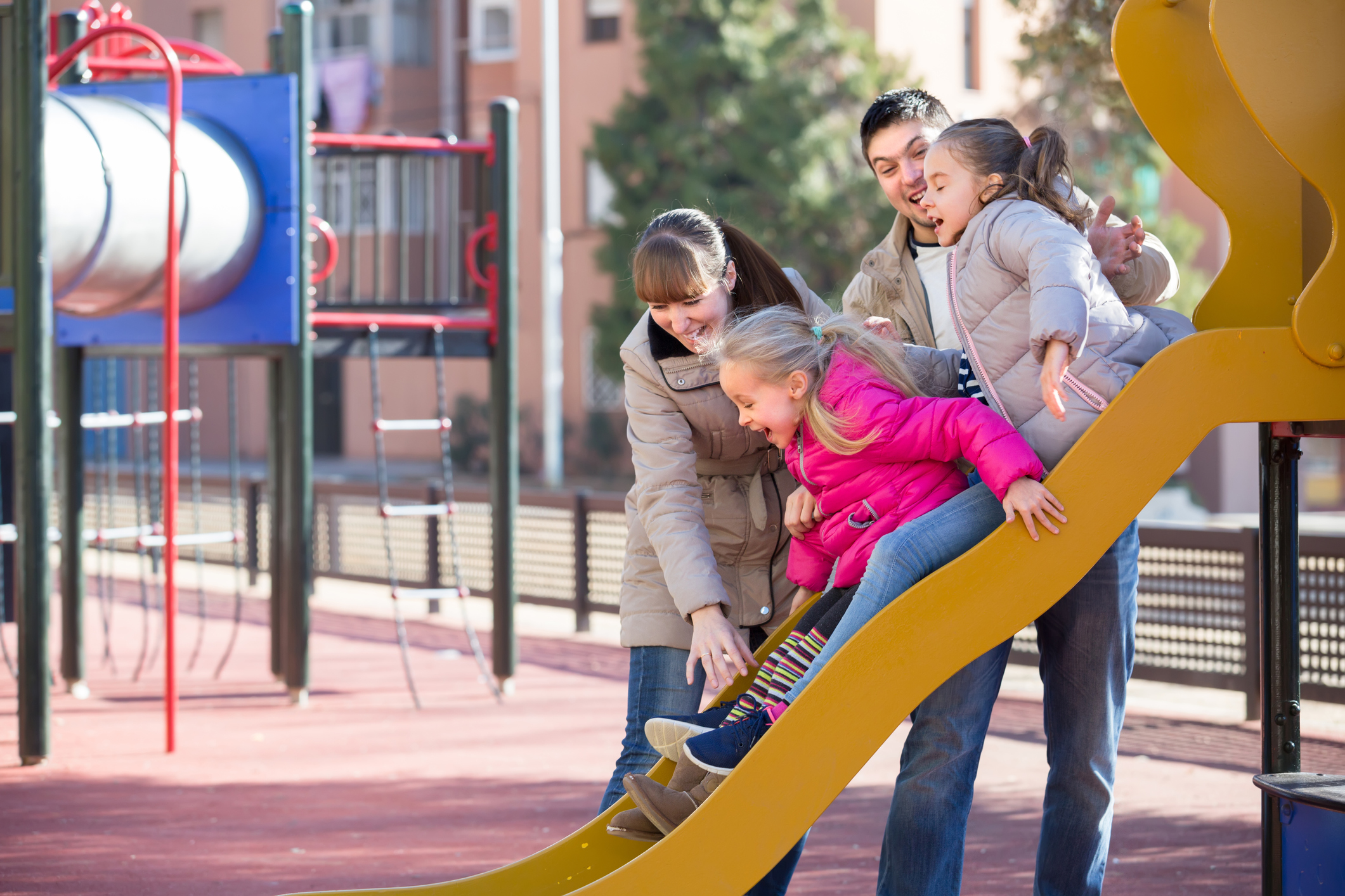 Man and woman helping two girls down a slide at a playground.