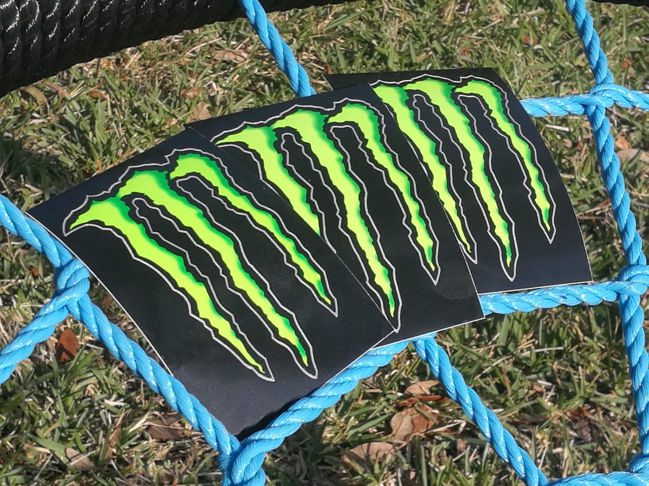 Three decals with Monster Energy logos resting on a bright blue mesh of ropes.