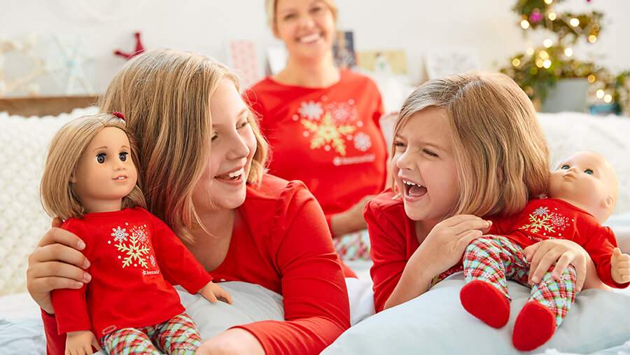 Two girls holding dolls in front of their mother, all three of them wearing matching red holiday pajamas, laughing and smiling on Christmas morning.