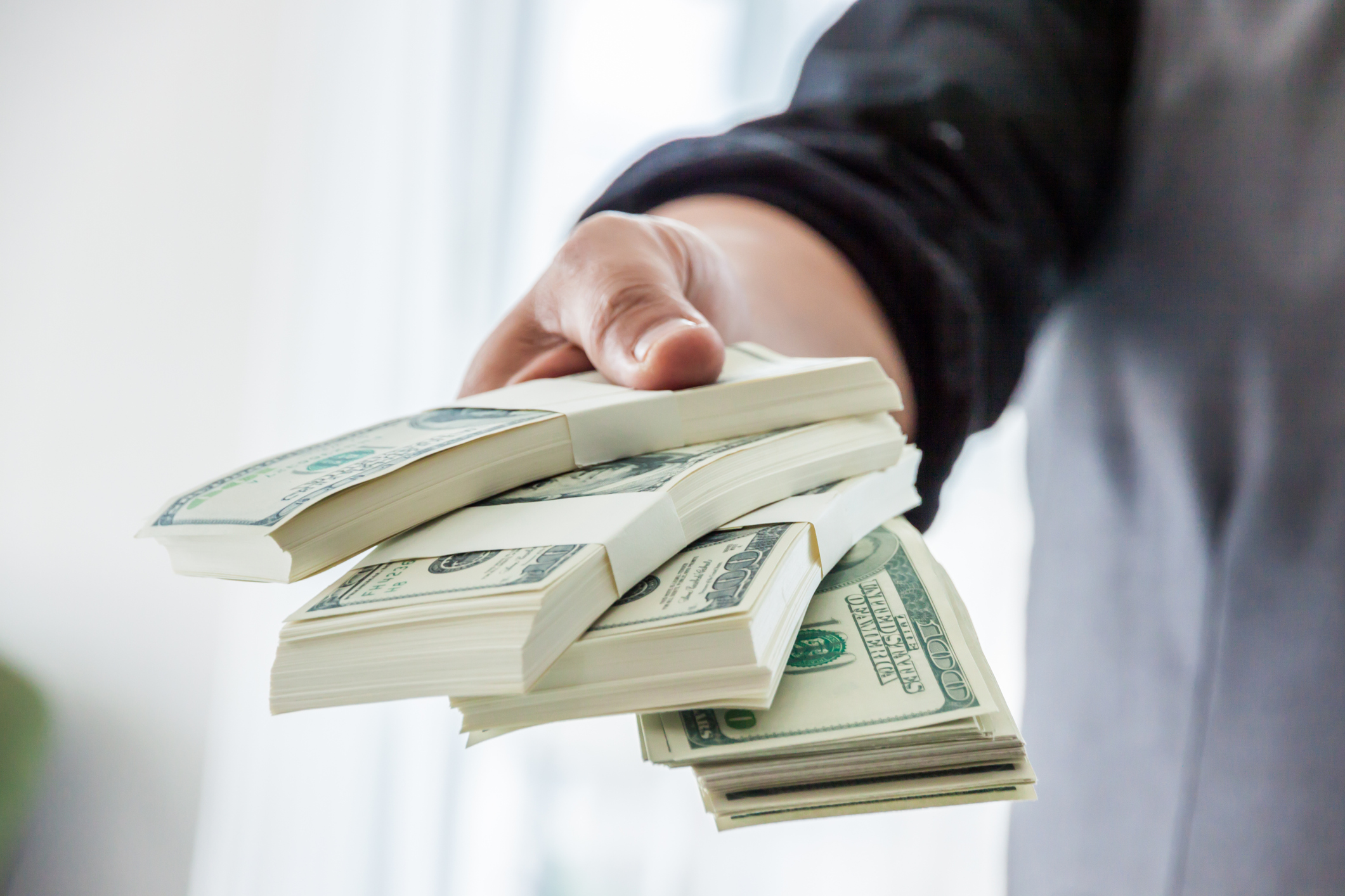 A hand reaching out holding stacks of $100 bills.