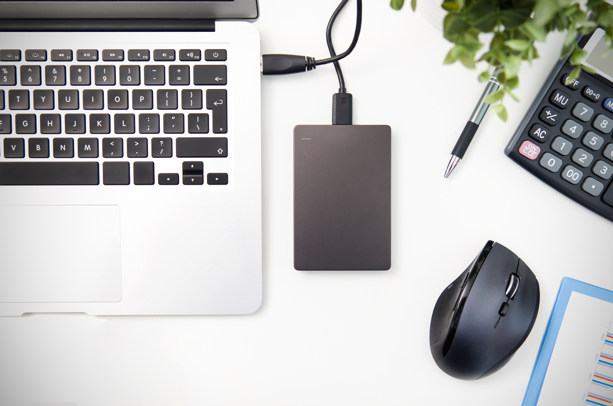 An external storage drive connected to a laptop with a pen, mouse, and calculator lying next to it.